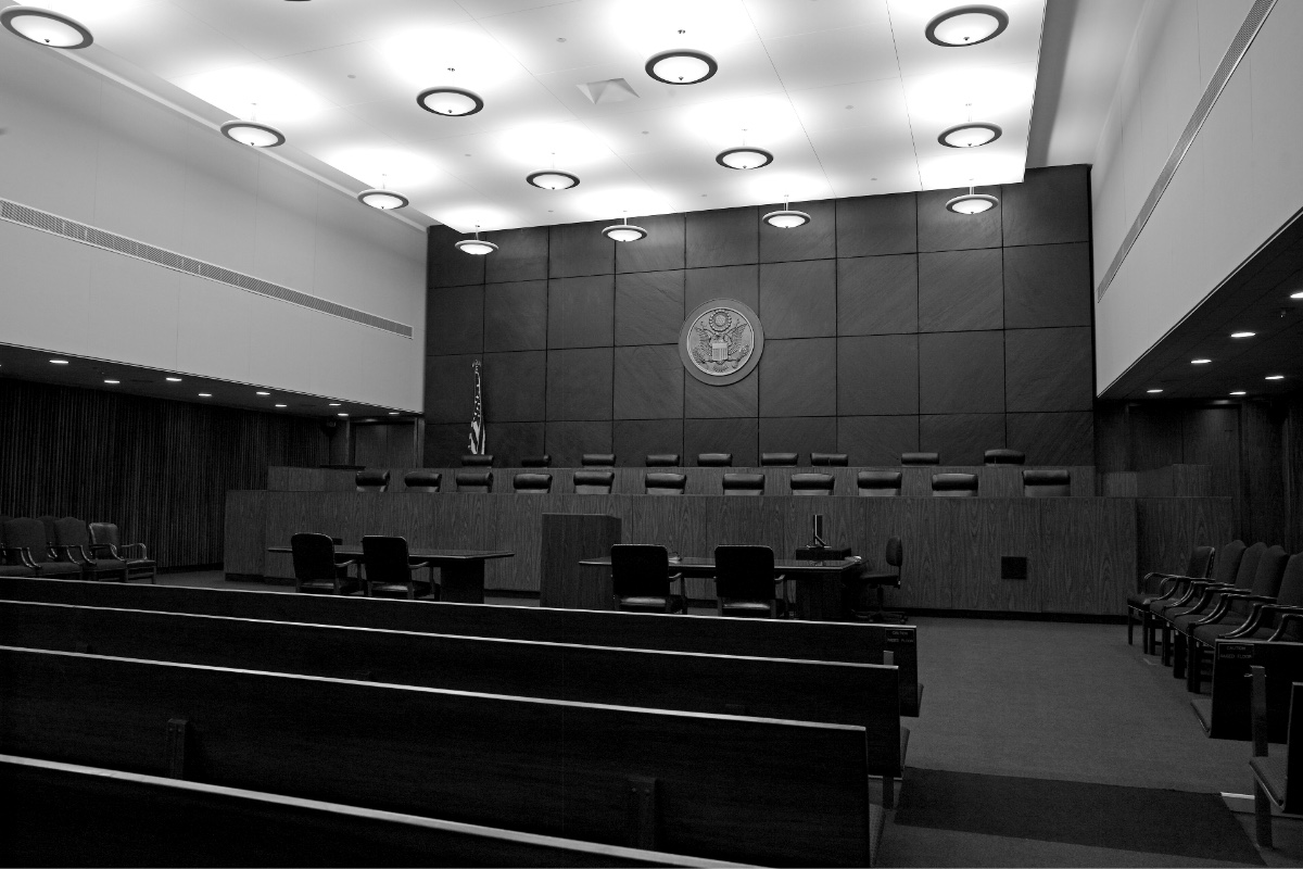 An empty federal courtroom with a long raised bench and a row of judges’ chairs beneath a dark panelled wall bearing a round seal, rows of wooden public benches in the foreground and round ceiling lights overhead, in black and white
