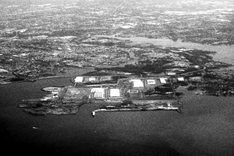 Sparrows Point in Baltimore County seen from the air, a flat peninsula of warehouses and cleared land reaching into the water, in high contrast black and white with coarse grain