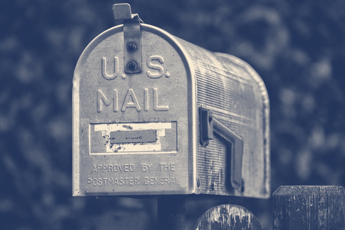 A weathered aluminium United States mail box with its red flag raised, on a fence post, in a slate and bone duotone