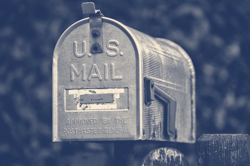 A weathered aluminium United States mail box with its red flag raised, on a fence post, in a slate and bone duotone