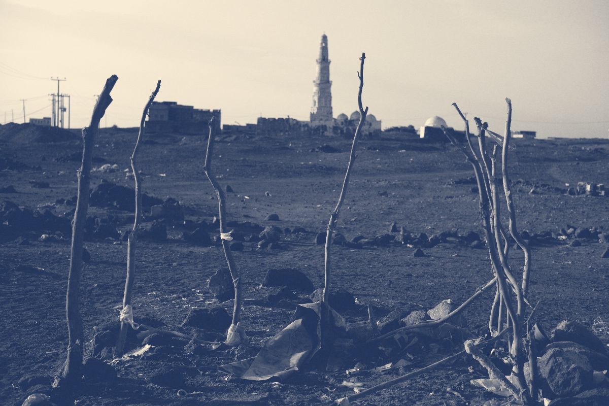 The minaret of the old mosque at Mokha rising beyond stony open ground, bare stakes and scattered litter in the foreground, in a slate and bone duotone