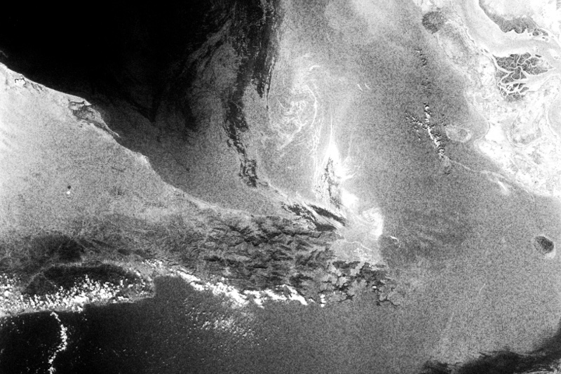 A satellite photograph of the Strait of Hormuz, the narrow channel between desert coastlines, in high contrast black and white with coarse grain