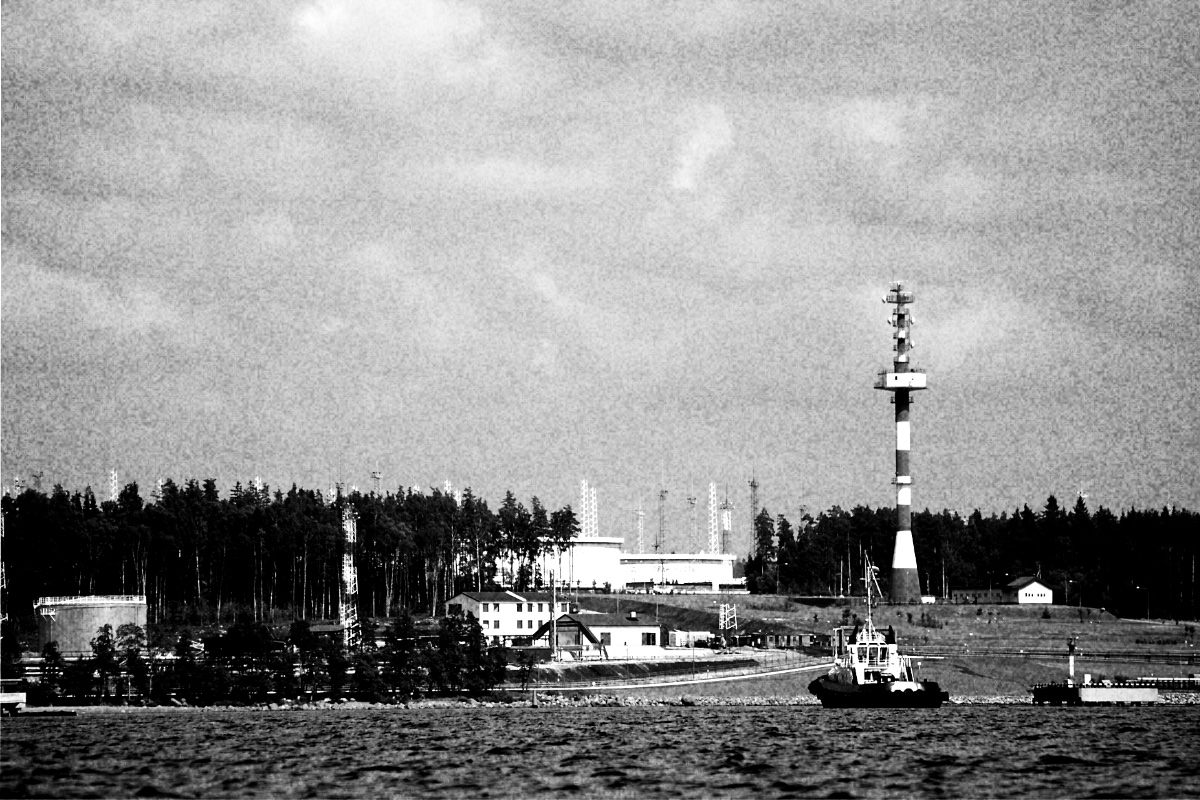The oil terminal at the Russian Baltic port of Primorsk seen from the water, a red and white striped lighthouse tower and low port buildings along a wooded shore, a tug in the foreground and storage tanks at the left, in grainy high-contrast black and white