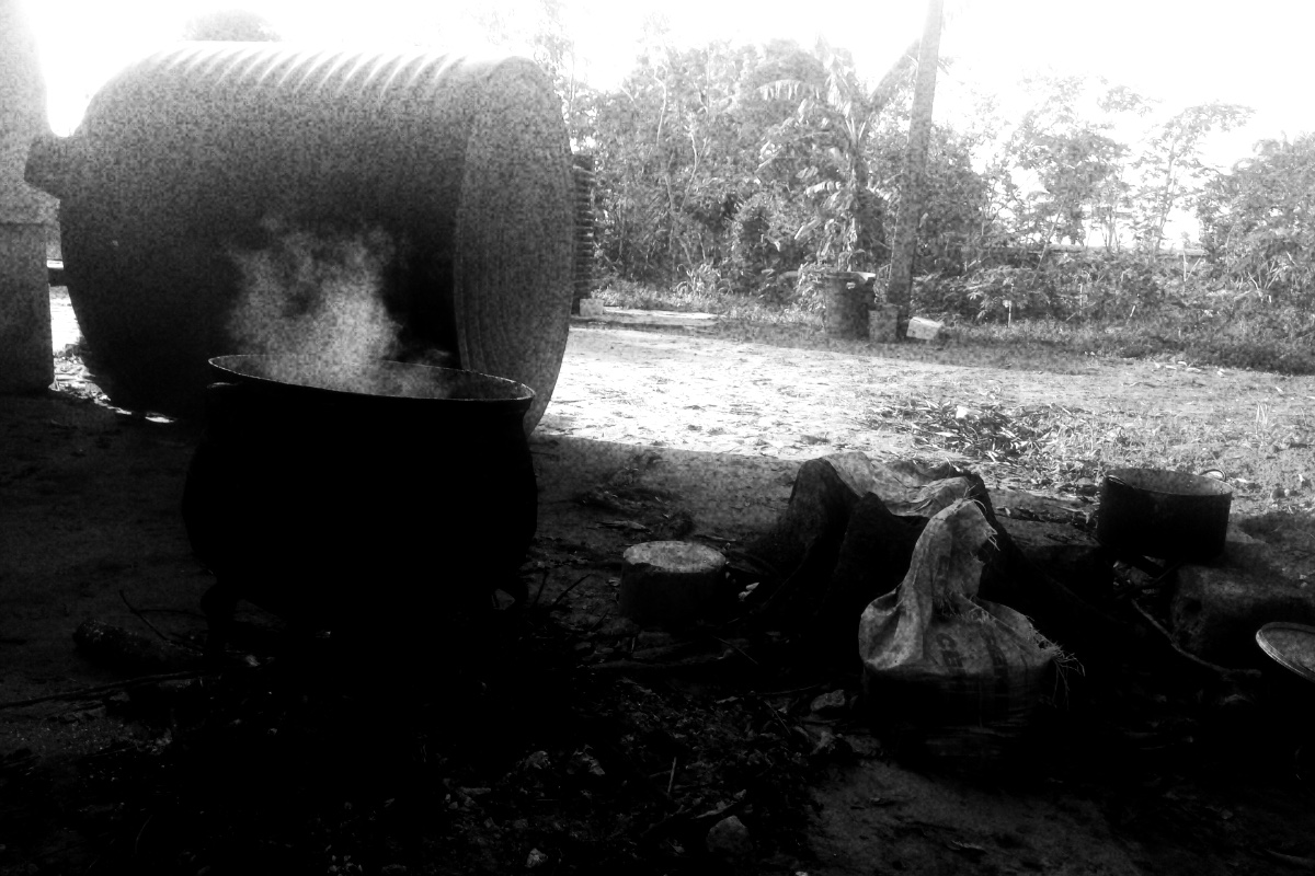 A black iron cooking pot steaming over a wood fire on bare ground outdoors, a large water tank and trees behind it, in grainy high-contrast black and white