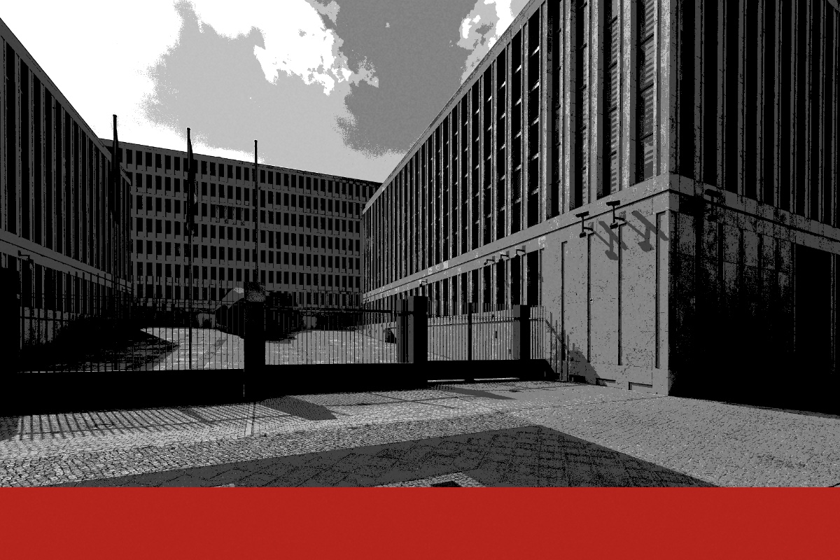 The gated forecourt of the Federal Intelligence Service headquarters in Berlin, long stone office blocks on either side and flagpoles behind the railings, in near black and white with a flat red band along the foot of the frame