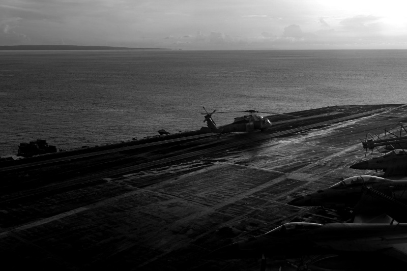 The empty flight deck of an aircraft carrier at dusk with a helicopter and parked aircraft, open sea and a distant coastline beyond, in plain black and white