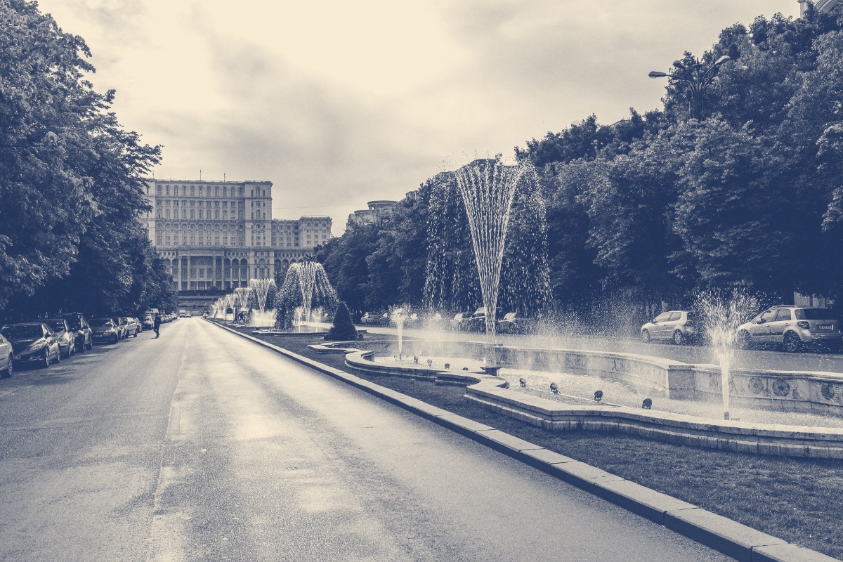Bulevardul Unirii in Bucharest seen along its central fountains towards the Palace of the Parliament at the far end, trees and parked cars lining the road, in a slate and bone duotone