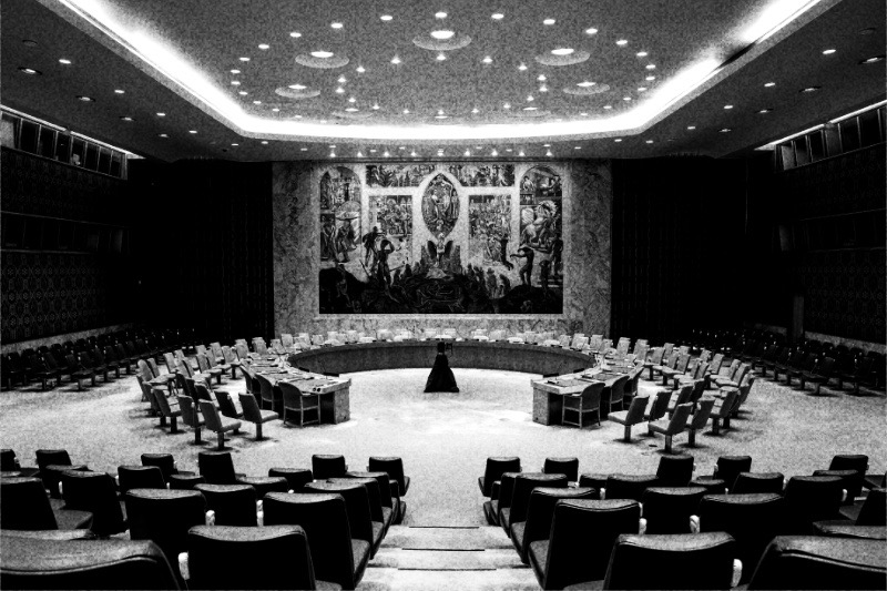 The empty Security Council chamber at UN headquarters in New York, its horseshoe table and rows of seats facing the large mural on the far wall, in high contrast black and white with coarse grain