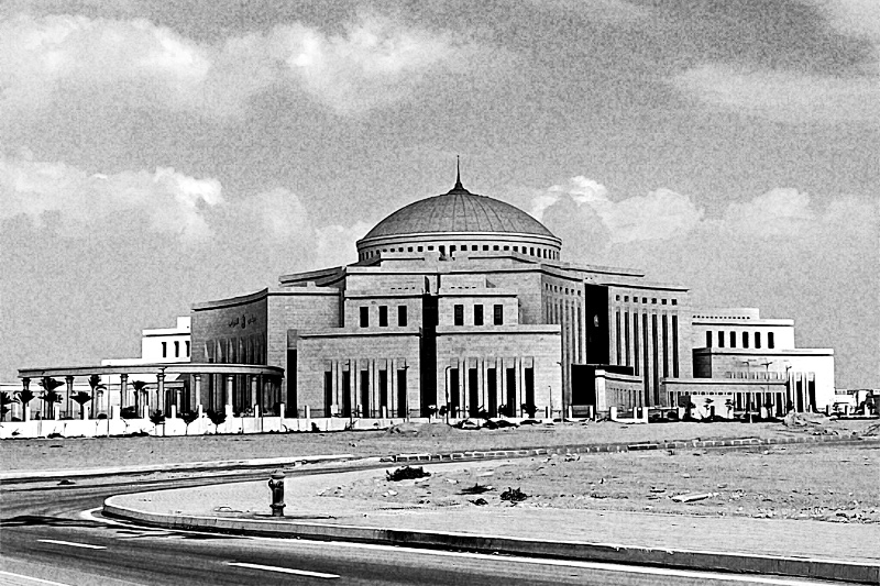 The domed parliament building in Egypt’s New Administrative Capital, a broad pale stone block with tall columns and a central dome, seen across an empty road and bare sandy ground under scattered cloud, in grainy high-contrast black and white