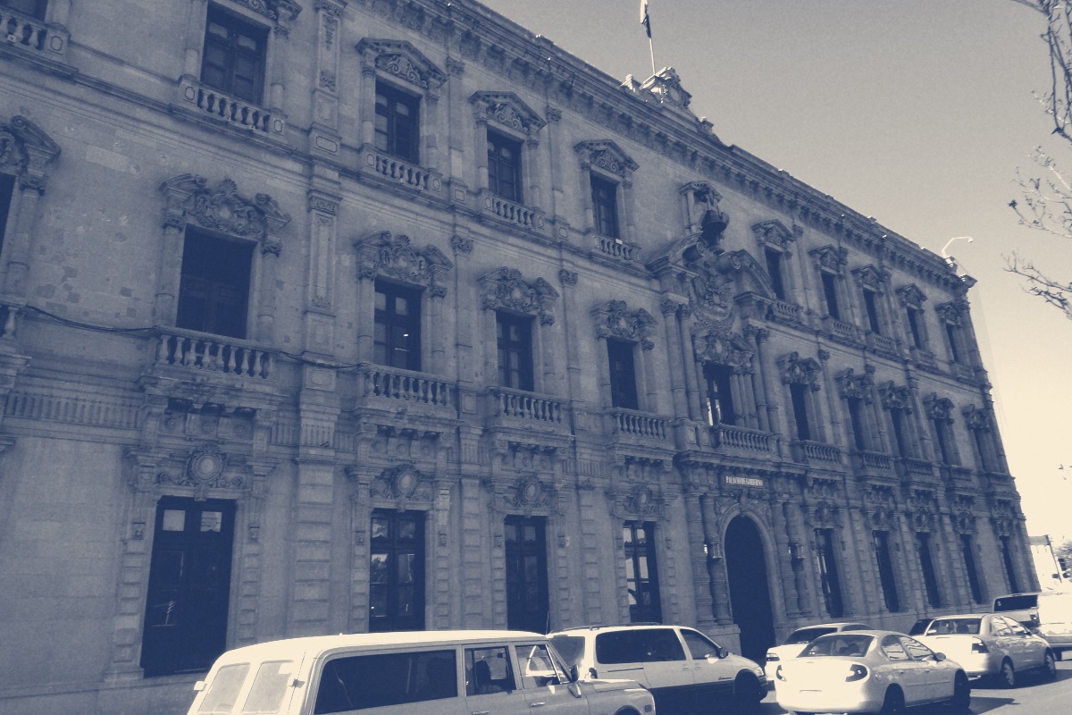 The carved stone facade of the Palacio de Gobierno in Chihuahua City, two storeys of tall windows and balconies under a flagpole, parked cars along the street in front, in a slate and bone duotone