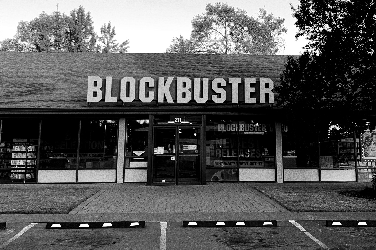 The storefront of the last Blockbuster video store, in Bend, Oregon, its name in large letters above a glass front with racks of films visible inside and an empty parking lot in front, in grainy high-contrast black and white