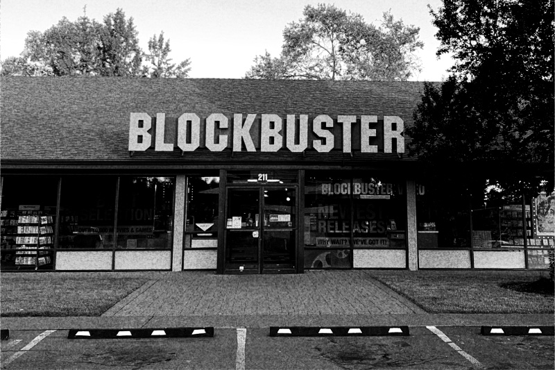 The storefront of the last Blockbuster video store, in Bend, Oregon, its name in large letters above a glass front with racks of films visible inside and an empty parking lot in front, in grainy high-contrast black and white