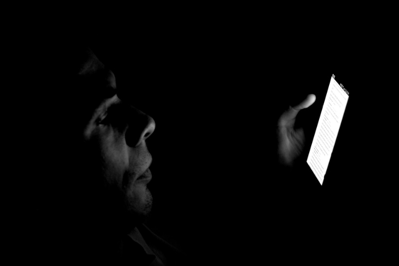 A man's face lit only by the glow of the smartphone he is reading in a dark room, in black and white