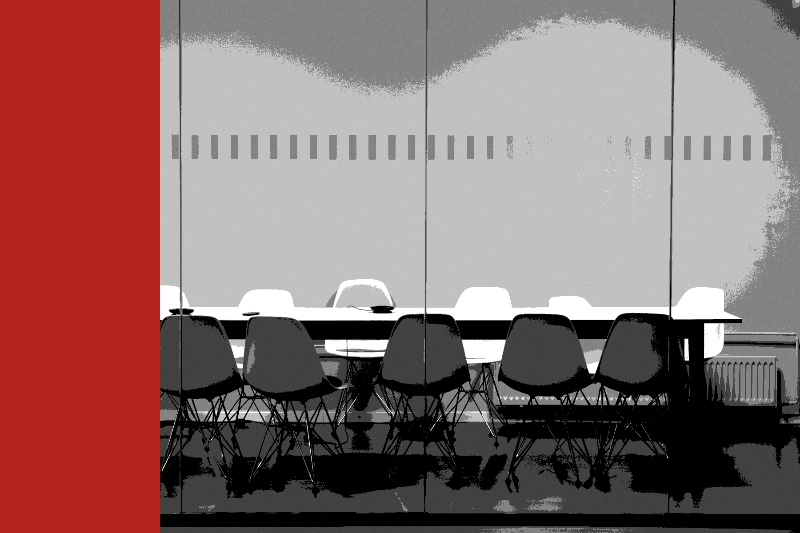 A row of empty chairs at a long meeting table seen through a glass partition, in near black and white with a red bar along the left edge