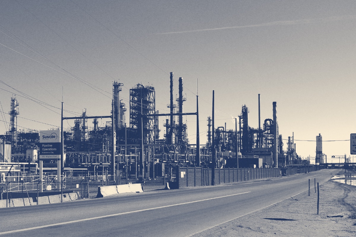 The Suncor Energy refinery in Commerce City, Colorado, towers and stacks along an empty road under a clear sky, in a slate and bone duotone