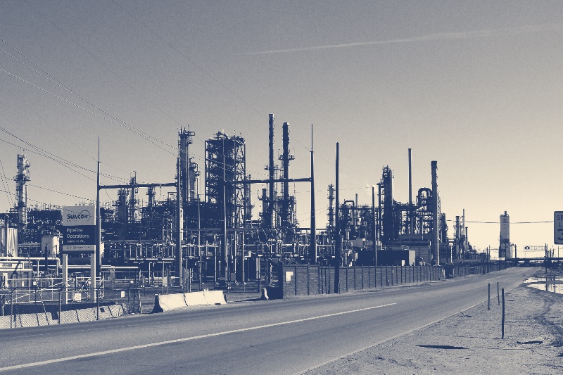 The Suncor Energy refinery in Commerce City, Colorado, towers and stacks along an empty road under a clear sky, in a slate and bone duotone