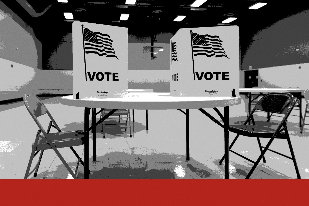 Two voting booths marked with the American flag and the word VOTE on a round table in an empty polling place, folding chairs beside it, in near black and white with a flat red band along the foot of the frame